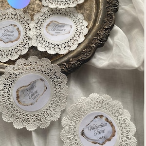May include: White lace doilies with printed name tags, displayed on a silver tray. Each tag features a name and table number within a decorative oval. The text "EDIT WITH CANVA" is visible in the upper left corner. The Etsy shop URL is visible at the bottom.