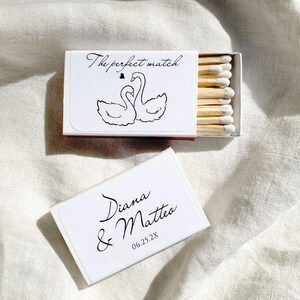 May include: White matchbox with the text "The perfect match" and a drawing of two swans. The box is open, revealing wooden matches with white tips. A second box is visible, with the names "Diana & Matteo" and the date "06.25.2X".