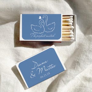 May include: Matchbox with a blue background and white text and swan illustration. The text reads "The perfect match" and "Diana & Matteo 06.25.2X". The matchbox is open, revealing wooden matches.