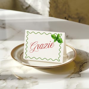 May include: A white place card with the word "Grazie" in red script, a green wavy border, and a basil leaf illustration. The card sits on a white plate with a gold rim, suitable for a dinner party.