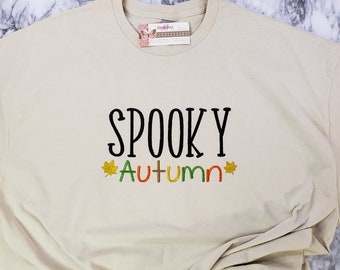 May include: A beige t-shirt with the words "SPOOKY AUTUMN" embroidered in black, green, orange, and yellow. The word "Autumn" features colorful leaf accents. The shirt is folded, and the background is a marble pattern.
