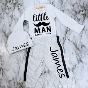 May include: A white baby outfit with a long-sleeved onesie, trousers, and a hat. The onesie has the text "little man" with a moustache graphic and an arrow. The trousers have a black stripe down the side and the name "James" written on the leg. The hat has the name "James" written on it.