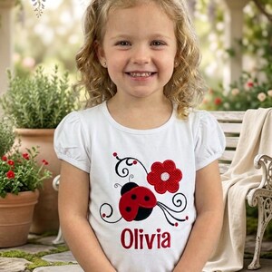 May include: A white short-sleeved t-shirt featuring a red and black ladybug graphic, a red flower, black swirling vine details, and the name Olivia embroidered in red lettering across the center of the chest area.