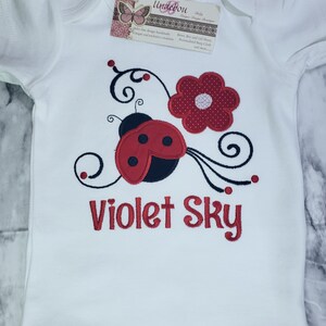 May include: A white short-sleeved baby bodysuit featuring an embroidered red and black ladybug, a red flower, and black swirling vines. The text Violet Sky is embroidered in red below the design. A rectangular brand tag is attached at the neckline.