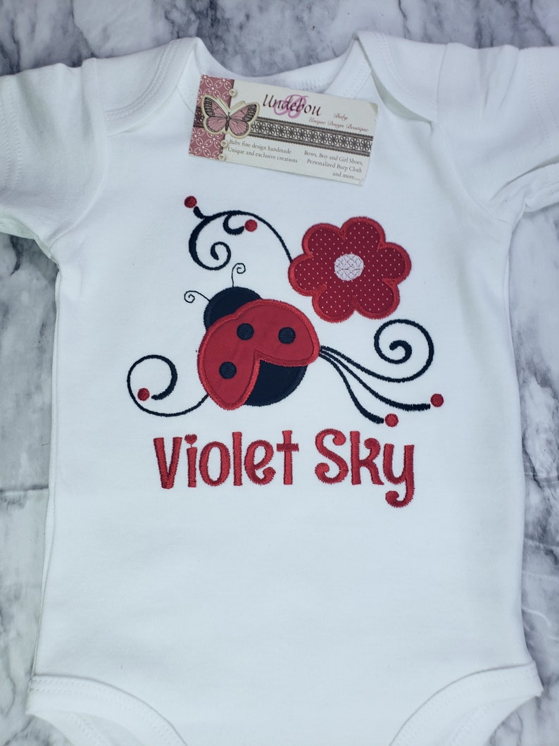 May include: A white short-sleeved baby bodysuit featuring an embroidered red and black ladybug, a red flower, and black swirling vines. The text Violet Sky is embroidered in red below the design. A rectangular brand tag is attached at the neckline.