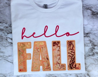 May include: White sweatshirt with the words "hello FALL" in red script and a fall-themed design. The word "hello" is in a cursive font, and the word "FALL" is in a block letter style with an orange and brown pattern.
