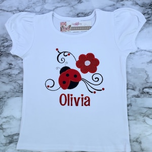 May include: A white short-sleeved t-shirt with puff sleeves featuring an embroidered red and black ladybug, a red flower, and black swirling vines. The name Olivia is embroidered in red below the design. A brand tag reads Undebou.