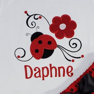 May include: A white shirt featuring an embroidered red and black ladybug, a red polka-dot flower, and black swirling vine accents. The name Daphne is embroidered in red lettering below the ladybug. A red and black polka-dot skirt is visible.
