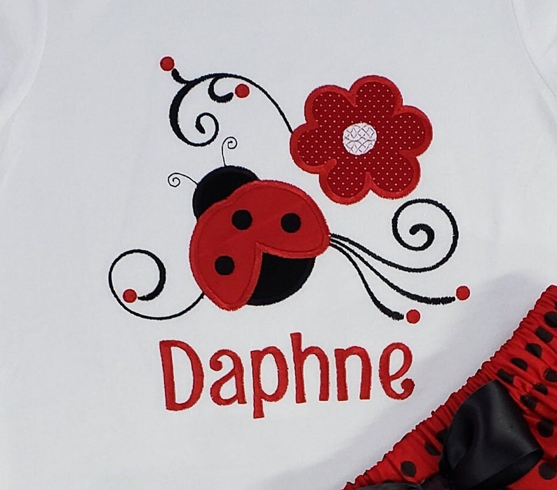 May include: A white shirt featuring an embroidered red and black ladybug, a red polka-dot flower, and black swirling vine accents. The name Daphne is embroidered in red lettering below the ladybug. A red and black polka-dot skirt is visible.
