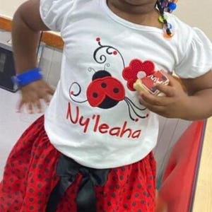 May include: A white short-sleeved t-shirt featuring a red ladybug graphic with black spots, a red flower, and the name Ny'leaha embroidered in red script. The shirt is paired with a red skirt featuring black polka dots and a black ribbon bow.