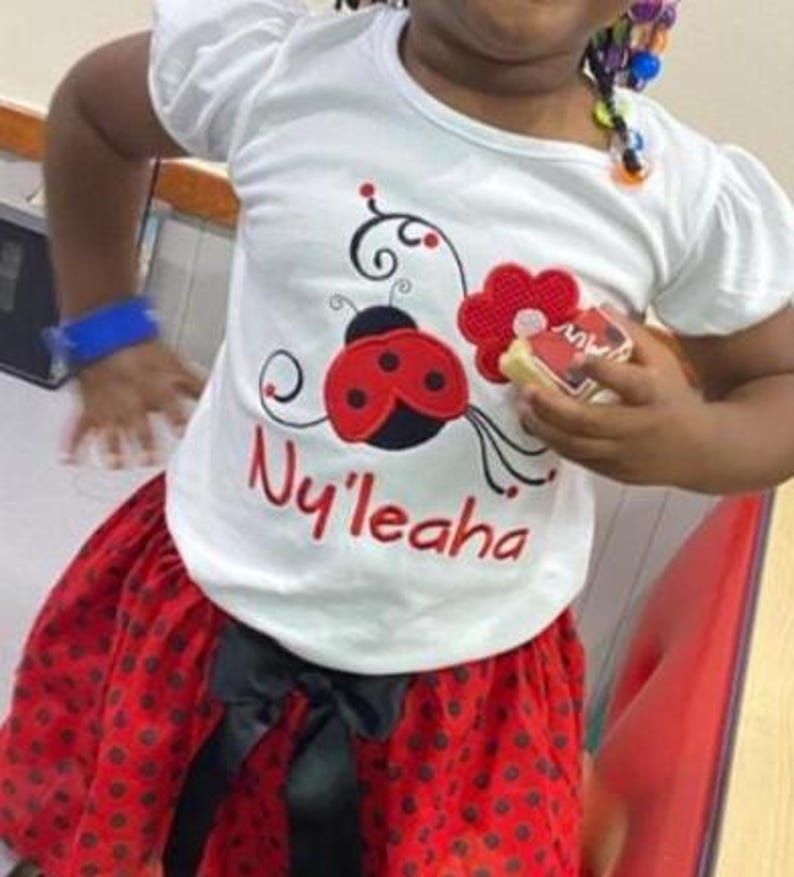 May include: A white short-sleeved t-shirt featuring a red ladybug graphic with black spots, a red flower, and the name Ny'leaha embroidered in red script. The shirt is paired with a red skirt featuring black polka dots and a black ribbon bow.