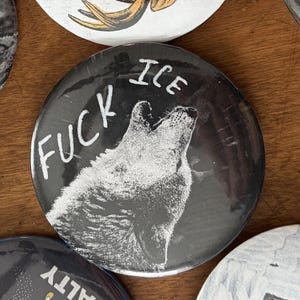 May include: A black button with a white illustration of a howling wolf. The button has the words "FUCK ICE" in white lettering. Other buttons with illustrations are visible in the background.