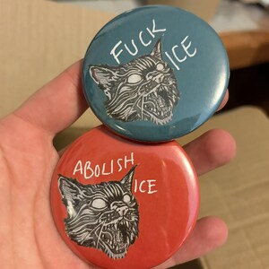 May include: Two round buttons with a cat graphic. One button is teal with the text "FUCK ICE". The other button is red with the text "ABOLISH ICE". The cat graphic is black and white.