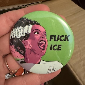 May include: A round, green button with the text "FUCK ICE" and a graphic of a woman with pink skin, black hair, and white streaks. The woman is screaming with her mouth open. The button is held in a hand.