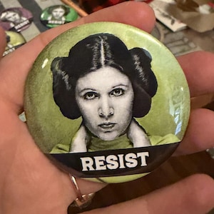 May include: A round button featuring a black and white image of a woman with a stern expression and a distinctive hairstyle. The word "RESIST" is printed in bold white letters across the bottom. The background is a light green.