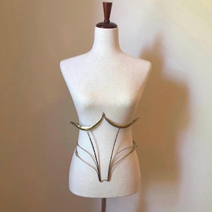 May include: A gold-toned metal body harness featuring two curved, crescent-shaped pieces at the chest area, connected by thin, structured wire framing that extends downward to form a cage-like design around the torso on a white mannequin.