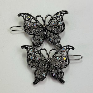 May include: Two dark gunmetal hair clips shaped like butterflies with intricate filigree cutouts and small iridescent rhinestones embedded throughout the wings. Each butterfly features a metal pin mechanism attached to the back for securing hair.