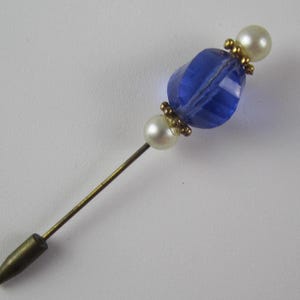 May include: A vintage hat pin with a faceted blue glass bead, two faux pearls, and gold-toned metal accents. The pin has a long, slender shaft and a pointed end. The design is elegant and suitable for adding a touch of vintage charm to any outfit.
