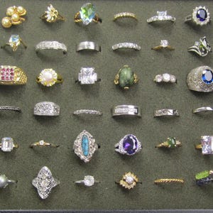 May include: A collection of thirty-five assorted rings displayed in a dark gray velvet jewelry tray. The rings feature various designs, including metal bands, gemstone settings, pearl accents, and clear stones in gold and silver-toned metals.