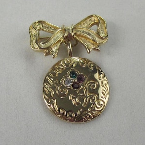 May include: A gold-toned brooch featuring a bow and a circular pendant. The pendant is adorned with an ornate floral design and four colorful gemstones. The brooch is a vintage accessory.