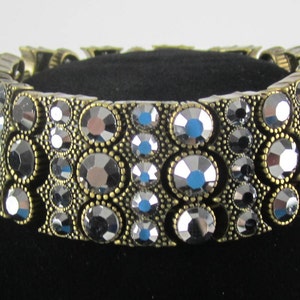 May include: A gold-toned bracelet with a row of large, faceted, black crystals. The bracelet is made of metal and has a hinged clasp.