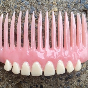Può includere: Un pettine di plastica rosa e bianco con una fila di denti in alto e una fila di denti bianchi in basso.