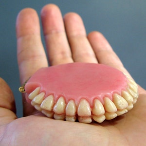 May include: A pink and white set of dentures with a gold clasp, held in a person's hand.