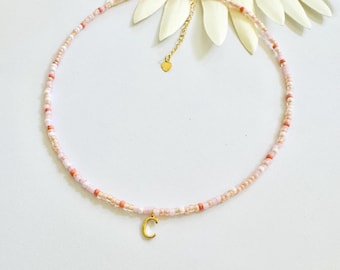 May include: A delicate beaded necklace in shades of pink, peach, and white. The necklace features a gold-colored initial pendant in the shape of the letter "C". The necklace is adjustable and has a gold-colored clasp.