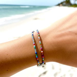 May include: A double-strand bracelet featuring small, multicolored glass seed beads in shades of blue, red, green, yellow, orange, and white, secured with a silver-toned metal clasp component at the base of the wrist.