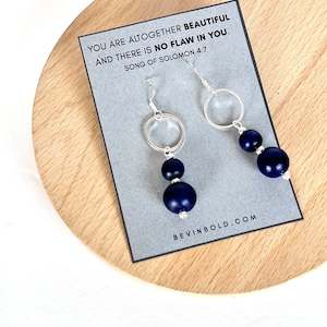 Puede incluir: Un par de aretes de aro de plata con dos cuentas de color azul oscuro que cuelgan de cada aro. Los aretes se muestran en una tarjeta con el texto "You are altogether beautiful and there is no flaw in you. Song of Solomon 4:7" y la dirección del sitio web "bevinbold.com".
