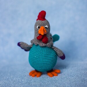 Puede incluir: Un juguete de gallo de crochet con cuerpo gris, cresta y barbilla rojas, pico naranja y plumas azules. El juguete está de pie sobre una superficie azul peluda.