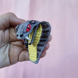 May include: A crocheted cobra snake with a yellow and black striped body, a gray head, and red eyes. The snake is in a striking pose with its mouth open.