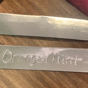 May include: Two silver metal plant tags. The top tag is blank. The bottom tag has the words "Orange Mint" written on it.