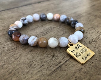 May include: A beaded bracelet with a gold charm that reads "be still and know" with the scripture reference "Psalm 46:10". The beads are a mix of white, brown, and gray.