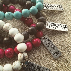 Peut inclure: Quatre bracelets avec des perles de différentes couleurs et des breloques argentées qui disent "Strong is Beautiful".