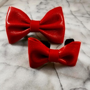 May include: Two red leather bow ties, one larger than the other, on a white marble surface.