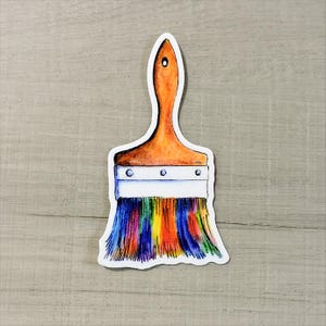 May include: A sticker of a paintbrush with a wooden handle and a white metal ferrule. The bristles are a vibrant mix of rainbow colors, including blue, yellow, orange, and purple. The sticker is on a light gray surface.