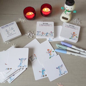 May include: A collection of white mini note cards featuring watercolor illustrations of snowmen with colorful scarves and hats. The cards are accompanied by Sharpie markers and candles, perfect for holiday greetings.
