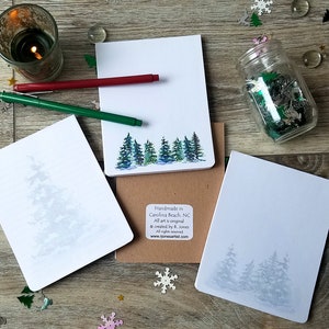 May include: Three white notepads with a watercolor design of pine trees. The top notepad has a blue and green watercolor design of pine trees. The bottom two notepads have a faint watercolor design of pine trees. The notepads are on a wooden surface with a jar of glitter, two markers, and other decorative items.
