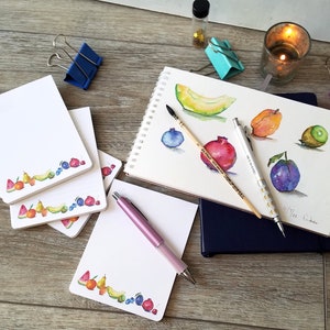 May include: A set of four white memo pads with a colorful fruit border. The pads are stacked on a wooden surface. A pink pen and a sketchbook with watercolor paintings of fruit are also visible.