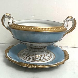 May include: A light blue and gold porcelain soup bowl with two handles, resting on a matching saucer. The bowl and saucer have gold trim and decorative detailing. The set is likely vintage and suitable for display or serving.