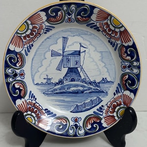 May include: A decorative plate featuring a windmill scene in blue and white, with a gold rim and floral border in red, yellow, and blue. The plate is displayed on a black stand, showcasing its intricate design and craftsmanship.