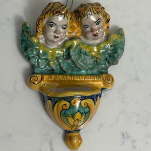May include: A ceramic wall pocket featuring two cherubic faces with yellow hair and rosy cheeks. The cherubs are surrounded by green and yellow wings. The pocket has a yellow and blue decorative design.