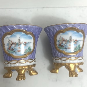 May include: Two matching lavender-colored porcelain vases with gold trim and feet. Each vase features a scenic painting of a harbor with boats and buildings. The vases have a scalloped rim and are likely decorative items.