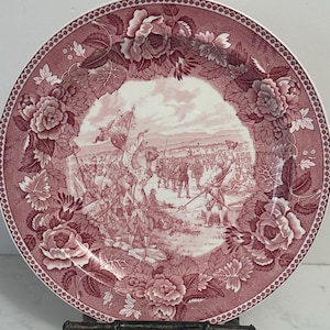 May include: A round ceramic plate featuring a deep red and white transferware pattern. The center depicts a historical military scene with soldiers and flags, while the wide border is decorated with detailed floral motifs and roses.