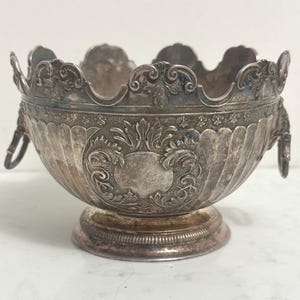 May include: An antique silver-colored bowl with ornate detailing. The bowl features a scalloped rim, decorative handles, and a pedestal base. Intricate floral and scrollwork patterns adorn the exterior, creating a vintage aesthetic. A classic piece for display or serving.