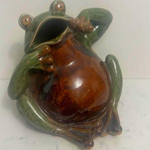 May include: A ceramic frog-shaped decorative container. The frog is primarily green with a brown belly and open mouth. The container has a glossy finish and is sitting on a white surface.