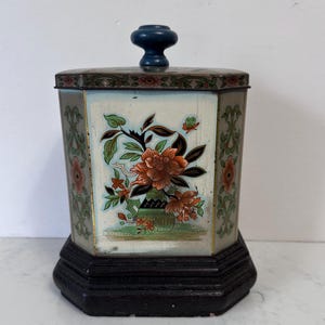 May include: An antique, hexagonal tin container with a dark wooden base and lid. The container features a floral design in orange, green, and black against a light blue background. The lid has a dark blue knob.