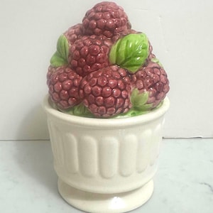 May include: A white ceramic container with a pedestal base, topped with a cluster of red raspberries and green leaves. The container has vertical grooves and a rounded rim, creating a decorative fruit-themed piece.