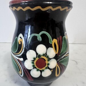 May include: A black ceramic vase with a wide, flared rim and a rounded body. The surface is decorated with hand-painted floral motifs featuring white petals, a dark red center, and green leaves, accented by yellow and white wavy line patterns.